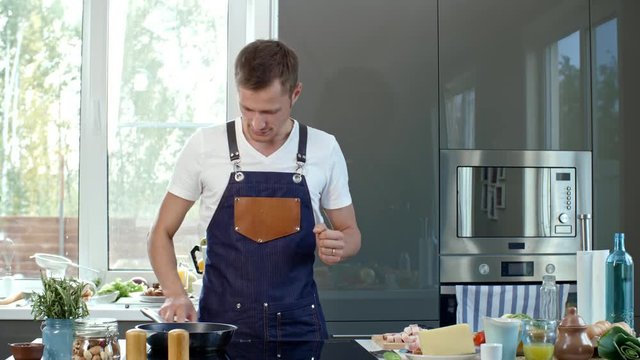 Medium shot of professional chef in apron standing at kitchen countertop and showing pan to camera while explaining something, then putting it on induction cooker and adding olive oil