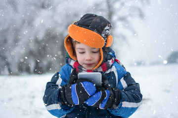 Portrait of little boy with mobile phone on snowy day in winter park.