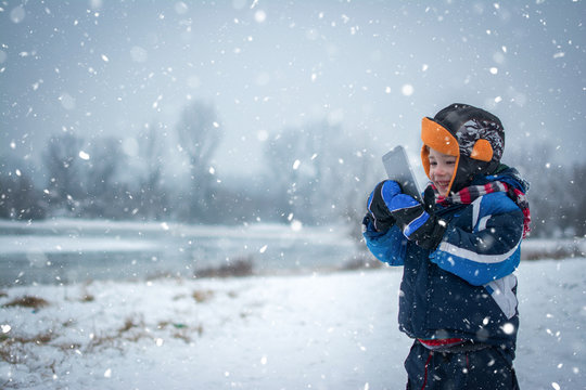 Smiling Little Boy With Mobile Phone On Snowy Day In Winter Park.