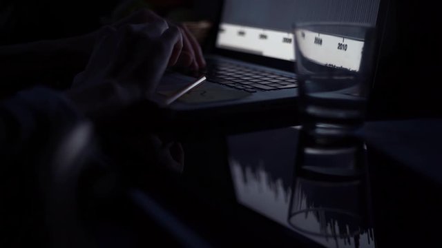 Young man using laptop in kitchen in evening. Glass of water and pencil on table