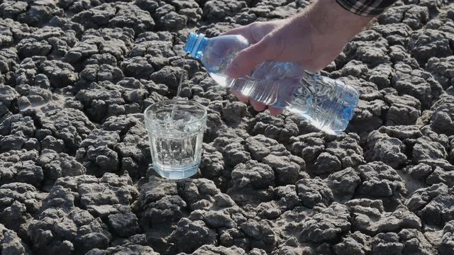 Human hand pouring clear drinking water to glass from bottle at dry cracked land