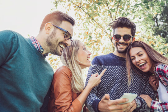 Image of four happy smiling young friends walking outdoors in the park holding smart phone.