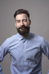 Fototapeta premium Childish young bearded hipster in formal wear winking with funny expression looking at camera against gray studio background. 