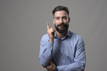Happy young bearded business man pointing finger upwards smiling and looking at camera with copyspace against gray studio background. 