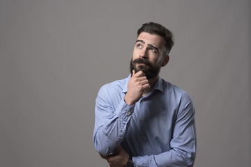 Young adult hipster business man thinking and looking up at copyspace while touching beard against...