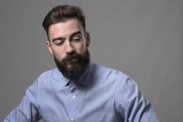 Young upset bearded adult hipster businessman looking down against gray studio background with copyspace