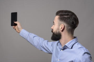 Profile view of young modern handsome bearded business man taking selfie photo with smartphone against gray studio background.