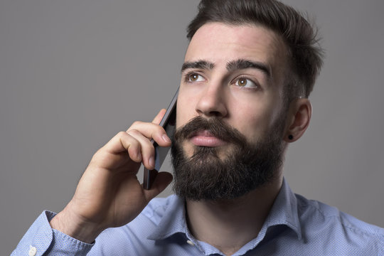 Close up portrait of young bearded business man talking on the mobile phone looking up at copyspace against gray studio background.