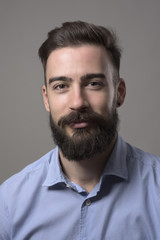 Obraz premium Moody bliska portret młodego brodatego mężczyzny, uśmiechając się i patrząc na kamery na szarym tle studio.