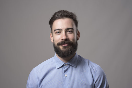 Horizontal  head and shoulder portrait of young bearded business man smiling at camera against gray studio background.