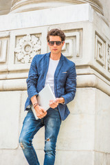 European college student studying in New York. Wearing blue blazer, white under shirt, jeans, sunglasses, holding laptop computer, a young guy standing by column on campus, looking at you, thinking..