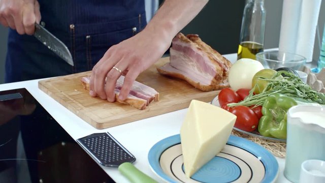 Handheld shot of unrecognizable cook chopping bacon on wooden board while cooking in kitchen; cheese and fresh vegetables lying on table