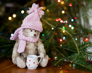 Old little teddy bear in knitted scarf and cap with soft focus on blurry bokeh christmas tree background