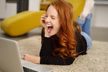 Fotobehang frau liegt auf dem fußboden im wohnzimmer und schaut lachend auf ihren laptop © contrastwerkstatt