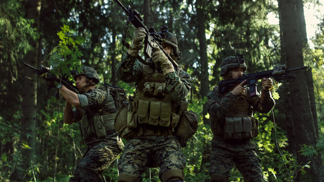 Three Fully Equipped Soldiers Wearing Camouflage Uniform Attacking Enemy, They're in Shooting Ready Stance, Aiming Rifles. Military Operation in Action, Squad Standing in Dense Forest.