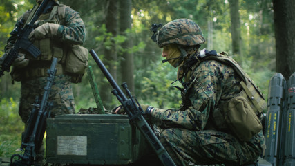 Military Staging Base, Chief Army Engineer Sitting on the Boxes of Ammunition Uses Army Grade Laptop Issued by Military Industrial Complex. Soldiers Prepare for Deployment.