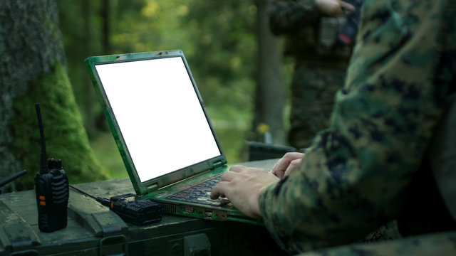 Close-up Soldier Uses Military Grade Laptop with White/Green Screen. In the Background Camouflaged Army Base in the Forest.