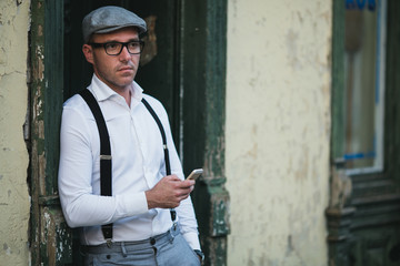 Fashionable retro dressed man with cap, suspenders and eyeglasses standing on city street and using smart phone.