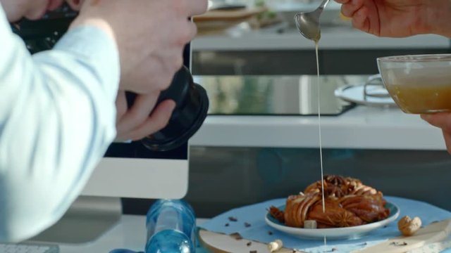 Handheld shot of unrecognizable man pouring syrup on Belgium waffles lying on plate on kitchen table as redhead man with digital camera taking still life photos