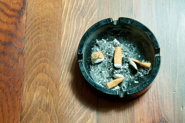 Dirty black ceramic ashtray full of cigaratte butts on the brown wooden table surface view from above