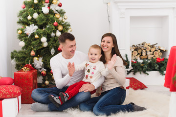 Happy young cheerful parents with cute little son. Child boy sitting in light room at home with decorated New Year tree and gift boxes. Christmas good mood. Family, love and holiday 2018 concept.