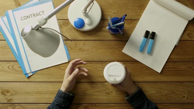 A man is sitting at the office desk in the phone and drink coffee. On the desk office supplies in blue and white tones.