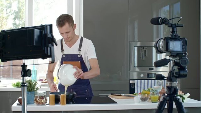 Professional chef in apron pouring creamy mixture into hot pan, then adding chopped spring onion and talking before digital camera while recording cooking show in kitchen