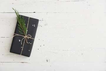 Top view on nice Christmas or birthday gift wrapped in black paper and decorated with cord and tree brunch on white wooden background.