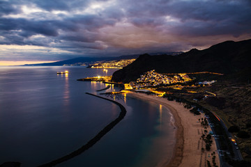 Dusk view of beautifully lit San Andres town with the capital city Santa Cruz de Tenerife in the background, Las Teresitas beach in the foreground and the North Atlantic Ocean to the left