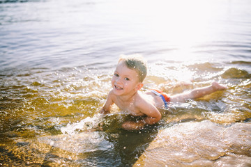 A Caucasian child of three years in red swimming trunks lies on his stomach in the water near the river bank of a sandy beach. Learns to swim with a smile