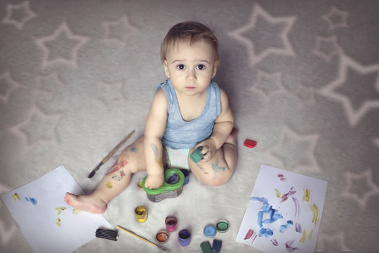 Cute boy covered in paint sitting on the floor among the items for creativity