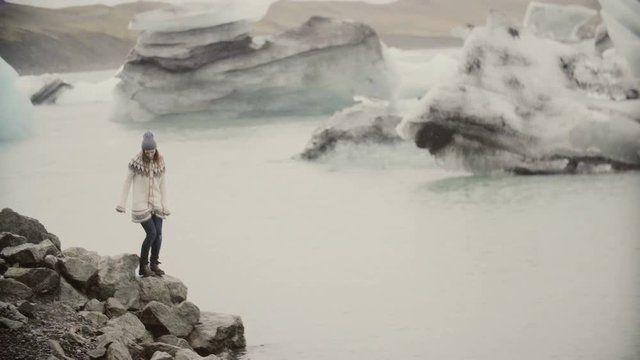 Young happy woman walking on the shore of ice lagoon. Tourist in lopapeysa exploring the sights of Iceland.
