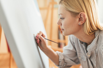 Fototapeta na wymiar Beautiful blonde woman artist drawing with a brush and pain in her art studio