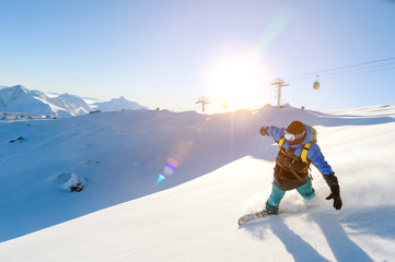 A snowboarder in a ski mask and a backpack is riding on a snow-covered slope leaving behind a snow powder against the blue sky and the setting sun