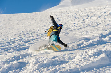 Freeride snowboarder rolls on a snow-covered slope leaving behind a snow powder against the blue sky