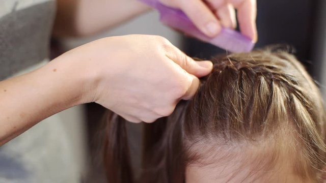 An attractive woman does her hairstyle for her little daughter. A woman plaits a pigtail of her daughter close-up.