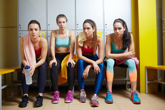 Four young women in activewear sitting in dressing-room before workout in gym
