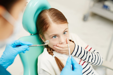 Scared little girl covering her mouth by hand while looking at dental tools for oral check-up held...