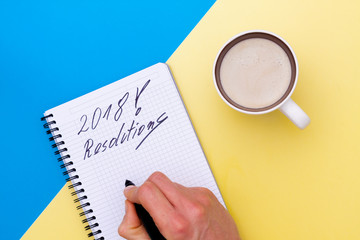 Top view on writing men hand in notebook and near cup of coffee, separated on yellow and blue paper background.. Hand write the new year resolutions.