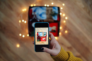 Woman taking photo of Christmas gift box with smartphone. Instagram photography blogging workshop concept. A girl hanging a phone taking a photo of present on wooden table.