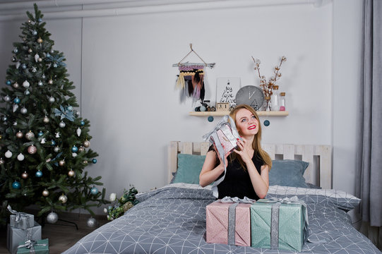 Cute blonde girl on black dress lying on bed with gifts boxes against new year tree at studio. Happy holidays.