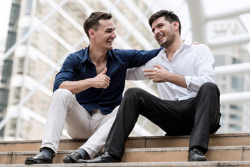 Two happy businessmen sitting down on concrete stairs shaking hands.