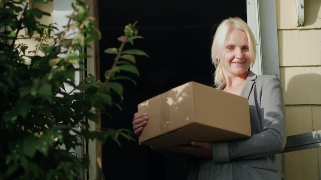 A woman with a parcel is standing on the threshold of her house. Looks at the camera, smiling. Delivery of parcels to the door