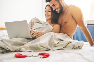Happy young couple lying on a bed with computer - Beautiful married couple watching role games sex video on laptop laughing together - People, sexual, technology concept - Soft focus on sheets