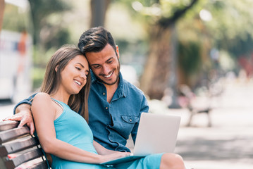Happy young couple using laptop computer sitting on a bench in city outdoor - Two lovers having fun spending time together to watch social network - Concept of love, people, addiction, technology
