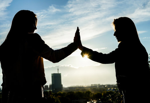 Silhouette of two young women giving high five having happy feelings standing on a hill at sunset in a park outdoor with city in the background - Concept of friendship, lifestyle, unity and travel