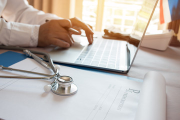 Doctor's working on laptop computer, writing prescription clipboard with record information paper folders on desk in hospital or clinic, Healthcare and medical concept. Focus on stethoscope