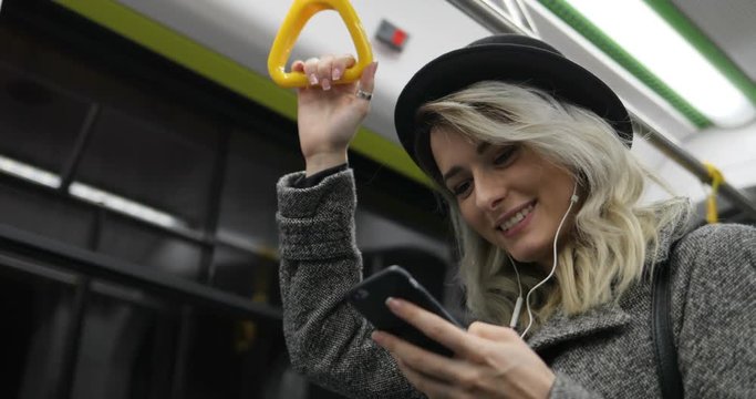 Portrait Of Cute Girl In Headphones Holds The Handrail, Listening To Music And Browsing On Mobile Phone In Public Transport. City Lights Background