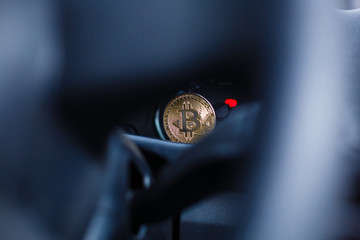 The physical coin is a gold bitcoin on the dashboard of the car next to the fuel consumption