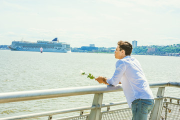 Fototapeta premium Man Missing You. Wearing white shirt, jeans, holding white rose, a guy standing by Hudson River in New York, opposite New Jersey, thinking, lost in thought. Concept of looking for love, friendship..